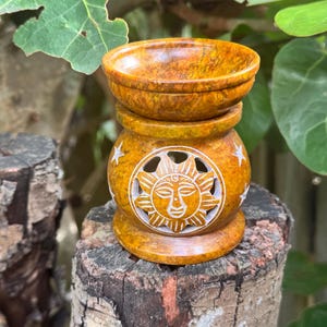 May include: A decorative, yellow-brown stone oil burner. The burner has a round bowl on top, a middle section with a carved sun design, and a round base. The sun design is white and has star accents. The oil burner is sitting on a tree stump.