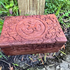 Puede incluir: Una caja rectangular de madera marrón rojizo con intrincadas tallas. La parte superior presenta un símbolo Om central dentro de un diseño circular, rodeado de patrones florales. La caja está colocada al aire libre entre vegetación.