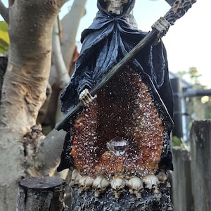 Puede incluir: Una figura esquelética con una túnica negra sostiene una guadaña y se encuentra frente a una cueva de cristal. La cueva está llena de cristales brillantes y rodeada por un anillo de cráneos.