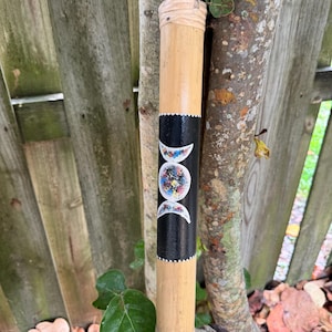 May include: A cylindrical bamboo incense holder with a black band featuring a colorful moon phase design. The holder is natural in color with a light-colored top and bottom. A black loop is attached to the top.