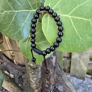 Pode incluir: Um bracelete de contas pretas com fecho de cordão preto. O bracelete é feito de contas redondas e polidas e é mostrado sobre uma superfície de madeira.
