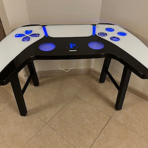 Op de afbeelding: Een zwart-witte gaming-tafel in de vorm van een PlayStation 5-controller met blauwe LED-verlichting. De tafel heeft een zwart blad en witte zijkanten met een PlayStation-logo in het midden.