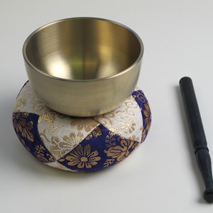 May include: A golden singing bowl rests on a patterned cushion with blue, white, and gold floral designs. A dark wooden striker lies beside the bowl. This image showcases a meditation and sound healing tool.