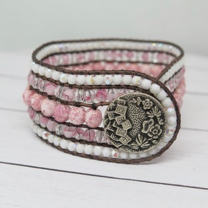 May include: A multi-strand bracelet with pink and white beads, brown leather, and a silver button with a floral design.