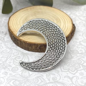 May include: A silver-toned crescent moon brooch featuring an intricate, openwork filigree lattice pattern throughout its curved body. The piece has a polished, smooth outer edge and a detailed, textured interior mesh design.