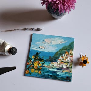 May include: A small oil painting depicting a coastal town with white buildings on a hillside overlooking a blue sea. The painting features a bright yellow citrus tree in the foreground and a blue sky with white clouds.