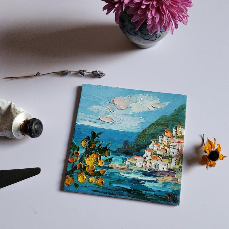 May include: A small oil painting depicting a coastal town with white buildings on a hillside overlooking a blue sea. The painting features a bright yellow citrus tree in the foreground and a blue sky with white clouds.