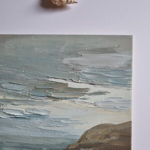 Pode incluir: Uma pintura a &oacute;leo retratando uma paisagem marinha com uma t&eacute;cnica de impasto texturizada. A obra de arte apresenta uma paleta de cores suaves de azuis, cinzas e marrons, com uma forma&ccedil;&atilde;o rochosa &agrave; direita. Uma concha &eacute; colocada acima da pintura.