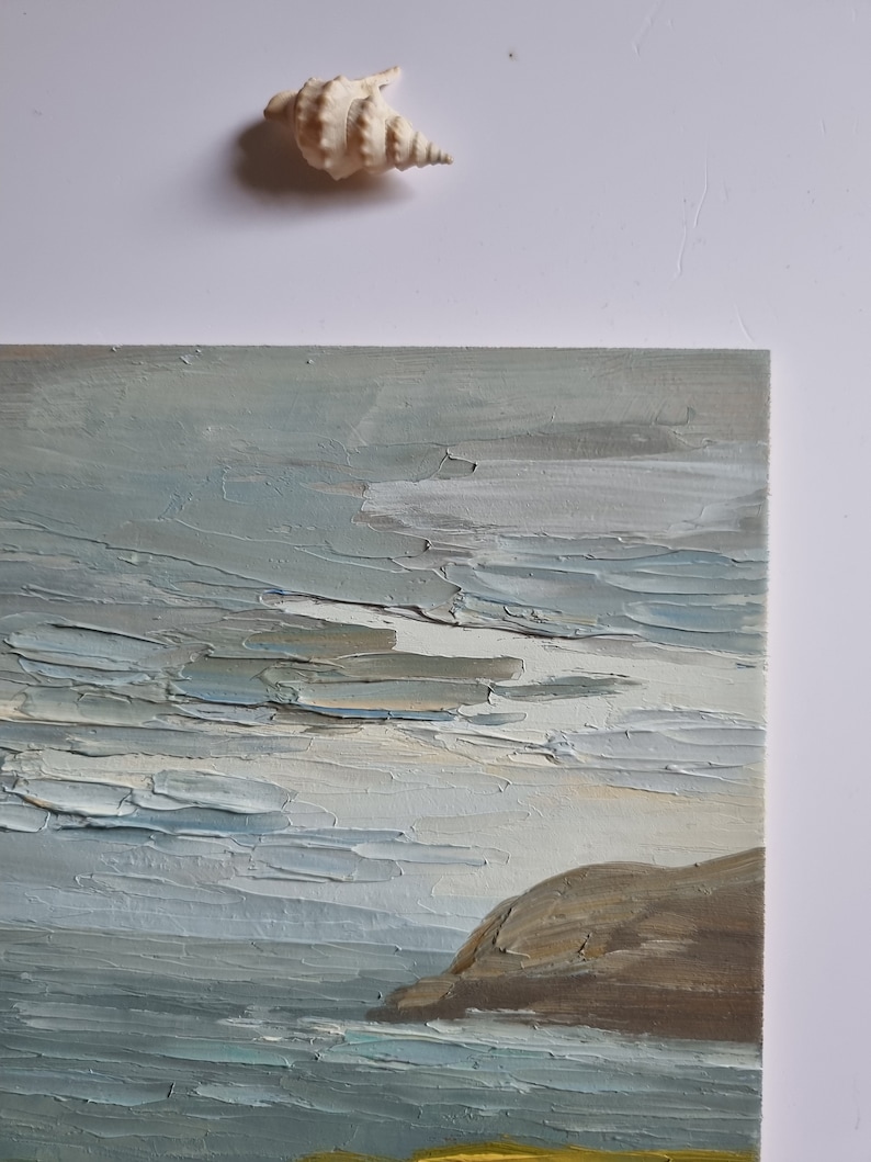 Pode incluir: Uma pintura a &oacute;leo retratando uma paisagem marinha com uma t&eacute;cnica de impasto texturizada. A obra de arte apresenta uma paleta de cores suaves de azuis, cinzas e marrons, com uma forma&ccedil;&atilde;o rochosa &agrave; direita. Uma concha &eacute; colocada acima da pintura.