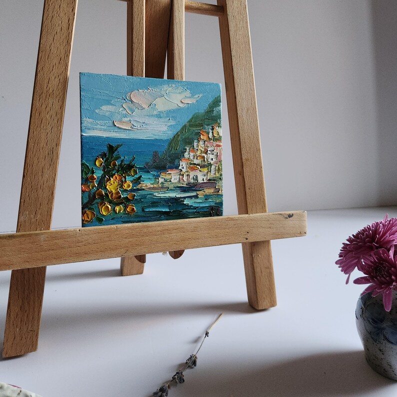 May include: A small painting on a wooden easel depicts a coastal town with white buildings on a hillside overlooking a blue sea. The painting is framed with a light blue border and features a small tree with yellow fruit in the foreground.