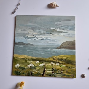 Pode incluir: Pintura a &oacute;leo de uma paisagem costeira com ovelhas pastando num campo verde. O c&eacute;u est&aacute; nublado com pinceladas texturizadas. Uma cerca de madeira est&aacute; em primeiro plano. Conchas e flores secas est&atilde;o dispostas ao redor da pintura.