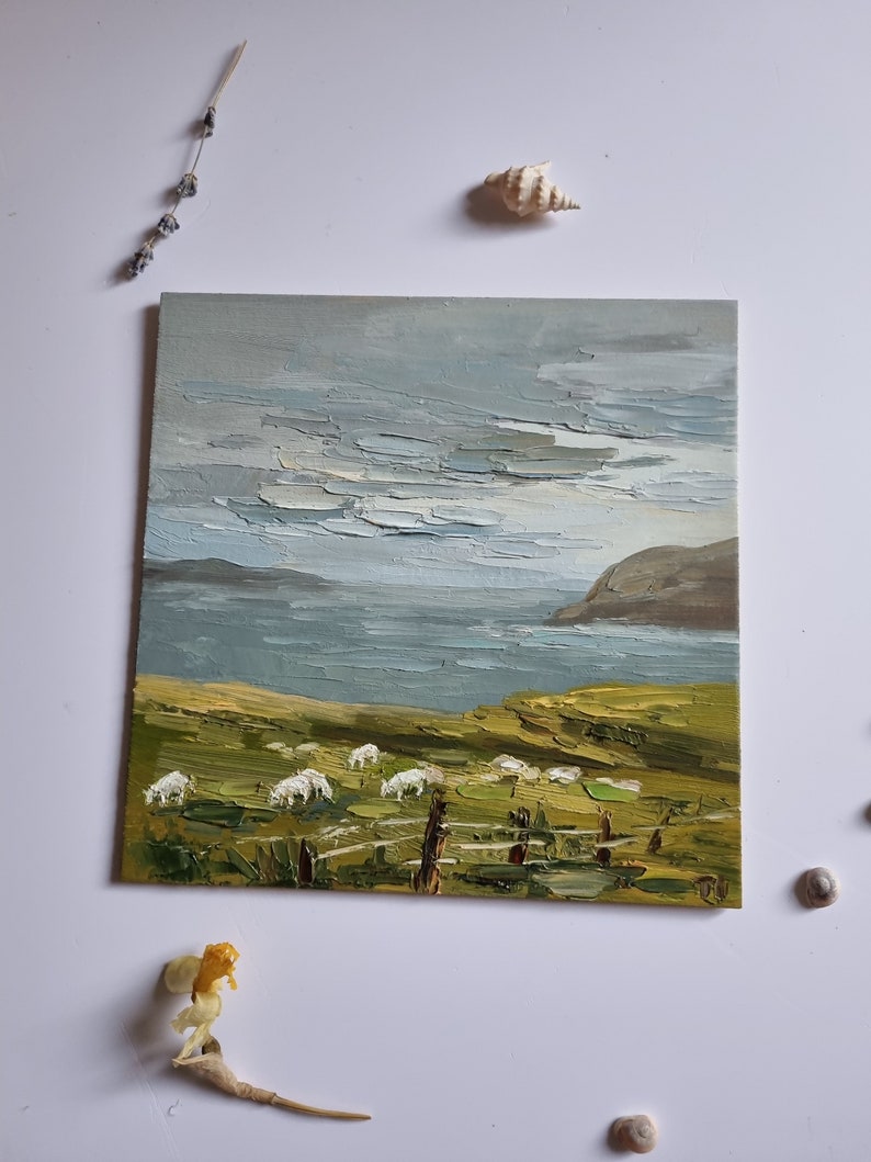 Pode incluir: Pintura a &oacute;leo de uma paisagem costeira com ovelhas pastando num campo verde. O c&eacute;u est&aacute; nublado com pinceladas texturizadas. Uma cerca de madeira est&aacute; em primeiro plano. Conchas e flores secas est&atilde;o dispostas ao redor da pintura.