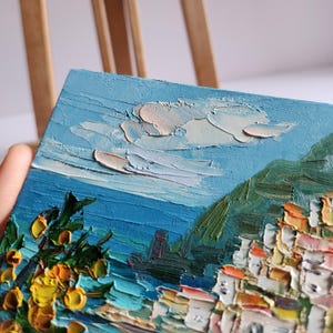 May include: A painting of a coastal town with white buildings, a green hillside, and a blue sea with a white cloud in the sky. The painting is done in a thick, impasto style.