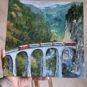May include: Oil painting of a red and white train crossing a stone bridge with arched supports. The bridge spans a valley with green trees and mountains in the background. The artwork is held by a person.