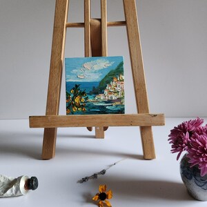 May include: A small painting on a wooden easel depicts a colorful seaside town with white buildings, green hills, and a blue ocean. The sky is light blue with white clouds. The painting is framed with a wooden frame.