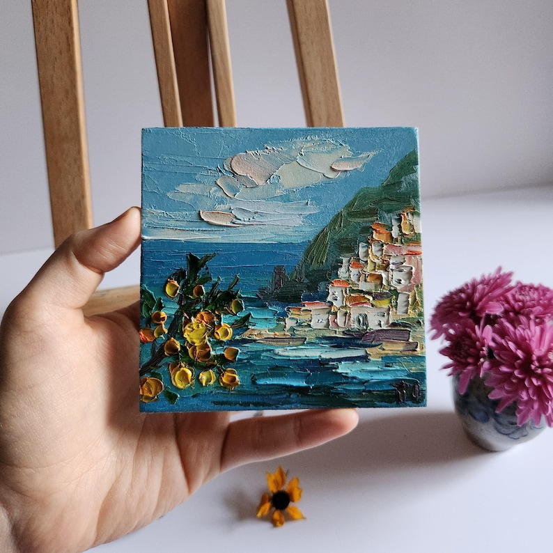 May include: A small oil painting on canvas depicting a coastal town with white buildings, a green hillside, and a blue sea with yellow flowers in the foreground.