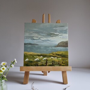 Pode incluir: Pintura a &oacute;leo sobre um pequeno cavalete de madeira que retrata uma paisagem costeira com ovelhas pastando num campo verde. O c&eacute;u est&aacute; nublado e o mar &eacute; vis&iacute;vel ao fundo. A pintura &eacute; exibida num pequeno cavalete de madeira.