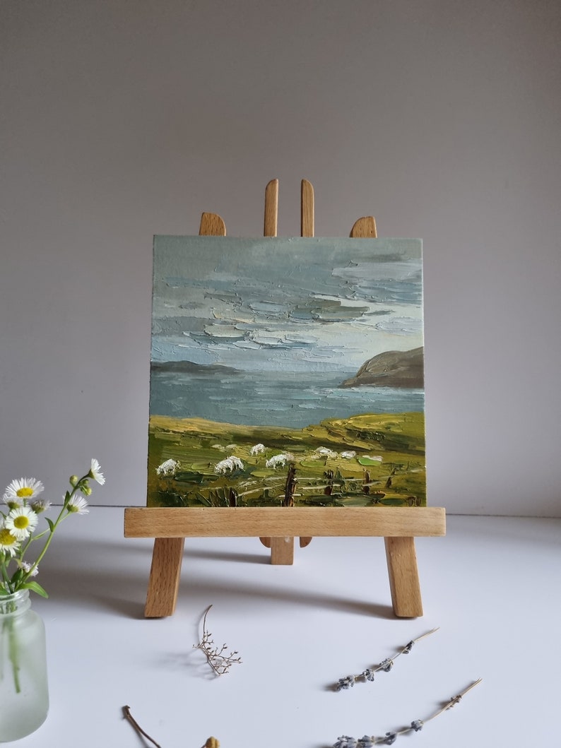 Pode incluir: Pintura a &oacute;leo sobre um pequeno cavalete de madeira que retrata uma paisagem costeira com ovelhas pastando num campo verde. O c&eacute;u est&aacute; nublado e o mar &eacute; vis&iacute;vel ao fundo. A pintura &eacute; exibida num pequeno cavalete de madeira.