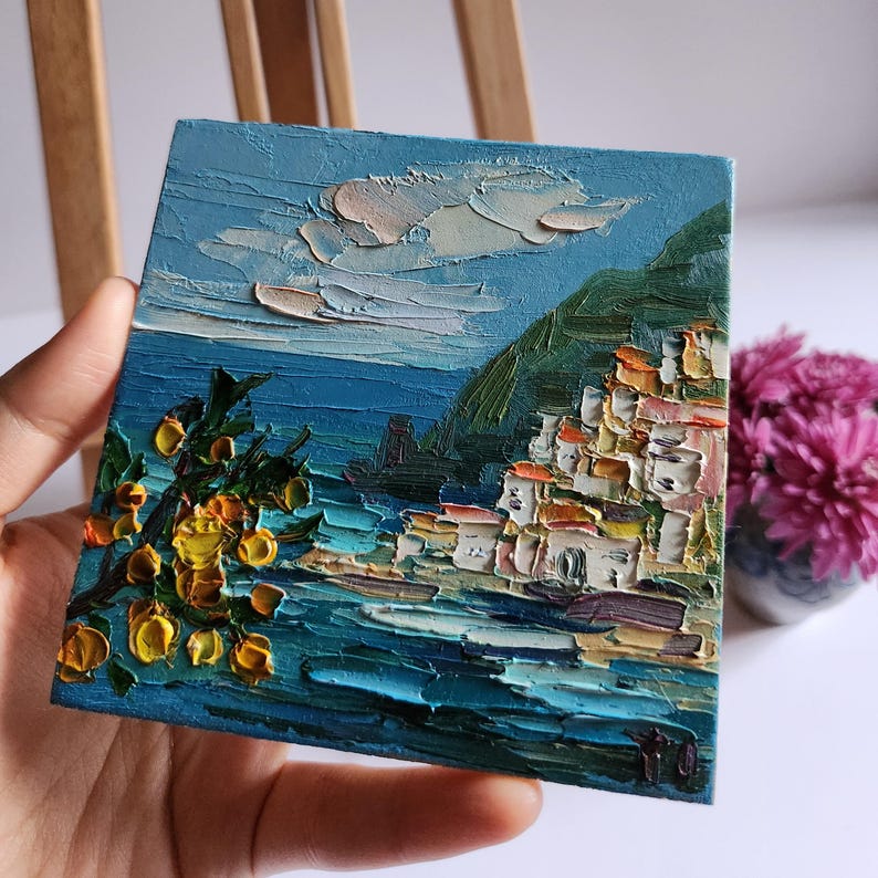 May include: A small oil painting depicting a coastal town with white buildings on a hillside overlooking a blue sea. The painting features a bright yellow flower bush in the foreground and a blue sky with white clouds.