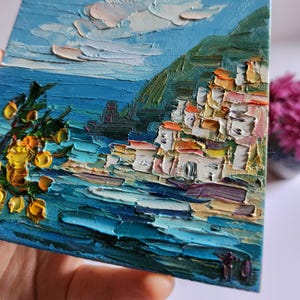 May include: A painting of a coastal town with colorful buildings on a hillside overlooking a blue ocean. The sky is a light blue with white clouds. There are yellow flowers in the foreground.
