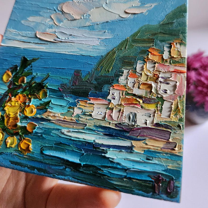 May include: A painting of a coastal town with colorful buildings on a hillside overlooking a blue ocean. The sky is a light blue with white clouds. There are yellow flowers in the foreground.
