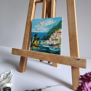 May include: A small painting on a wooden easel depicts a colorful seaside town with white buildings, a green hillside, and a blue sea. The sky is a light blue with white clouds. There are yellow flowers in the foreground.