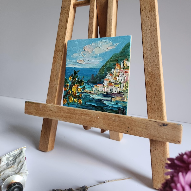 May include: A small painting on a wooden easel depicts a colorful seaside town with white buildings, a green hillside, and a blue sea. The sky is a light blue with white clouds. There are yellow flowers in the foreground.