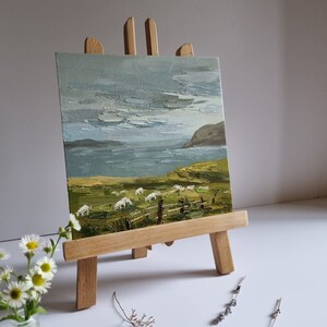 Pode incluir: Uma pequena pintura a &oacute;leo sobre um cavalete de madeira retratando uma paisagem costeira com ovelhas pastando em um campo verde. O c&eacute;u &eacute; cinza e azul, e o mar &eacute; vis&iacute;vel ao fundo. A pintura &eacute; exibida em um pequeno cavalete de madeira.