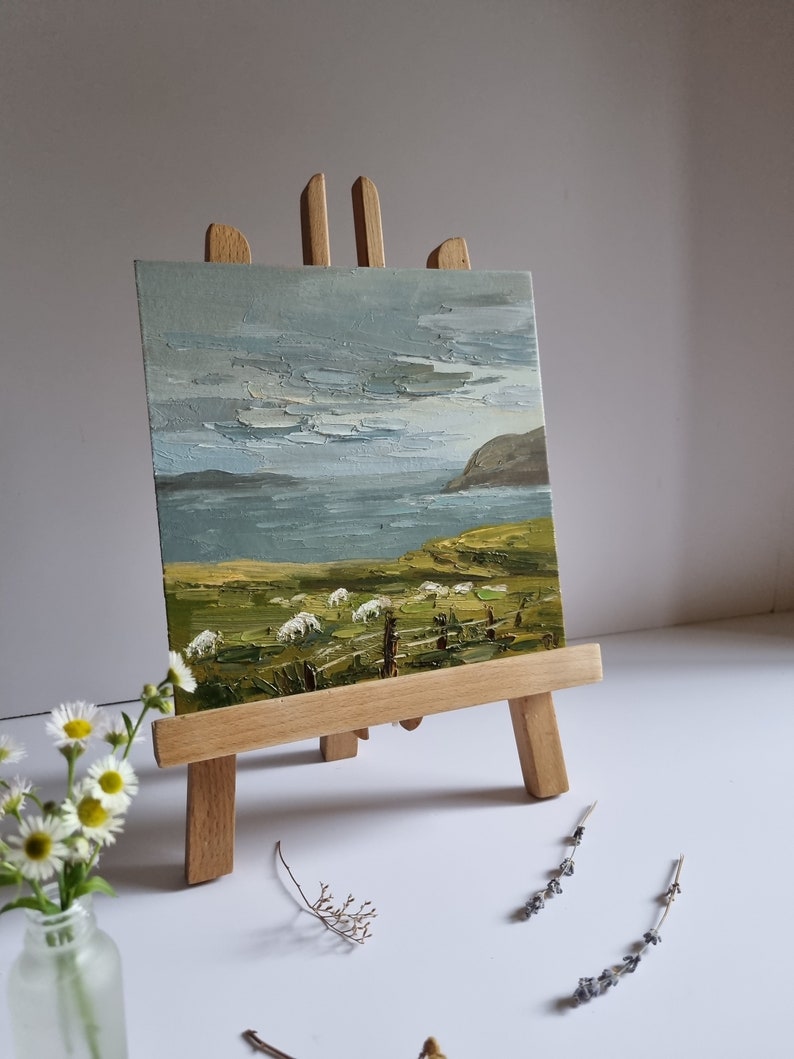 Pode incluir: Uma pequena pintura a &oacute;leo sobre um cavalete de madeira retratando uma paisagem costeira com ovelhas pastando em um campo verde. O c&eacute;u &eacute; cinza e azul, e o mar &eacute; vis&iacute;vel ao fundo. A pintura &eacute; exibida em um pequeno cavalete de madeira.