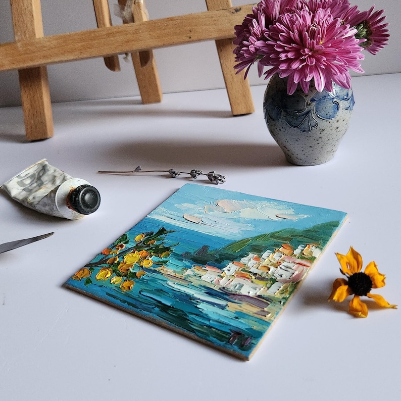 May include: A small painting of a coastal town with white buildings, a green hillside, and a blue sea. The painting is on a wooden easel and there are flowers in a vase in the background.