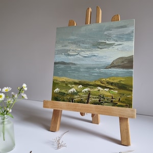 Pode incluir: Pintura a &oacute;leo sobre um cavalete de madeira que retrata uma paisagem costeira com ovelhas pastando num campo verde. O c&eacute;u &eacute; azul p&aacute;lido com pinceladas texturizadas e o mar &eacute; azul escuro. Um pequeno vaso de flores brancas &agrave; esquerda.