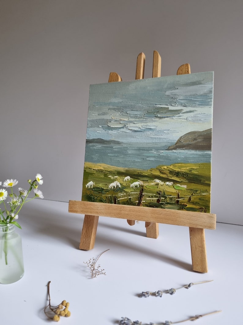 Pode incluir: Pintura a &oacute;leo sobre um cavalete de madeira que retrata uma paisagem costeira com ovelhas pastando num campo verde. O c&eacute;u &eacute; azul p&aacute;lido com pinceladas texturizadas e o mar &eacute; azul escuro. Um pequeno vaso de flores brancas &agrave; esquerda.