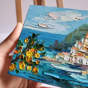 May include: A small oil painting depicting a coastal scene with a village nestled on a hillside. The painting features a bright blue sea, a green hillside, and a yellow citrus tree in the foreground.