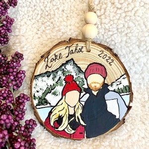May include: A wooden ornament with a painted scene of a couple in winter clothing standing in front of a mountain with the text "Lake Tahoe 2022" on the wood.