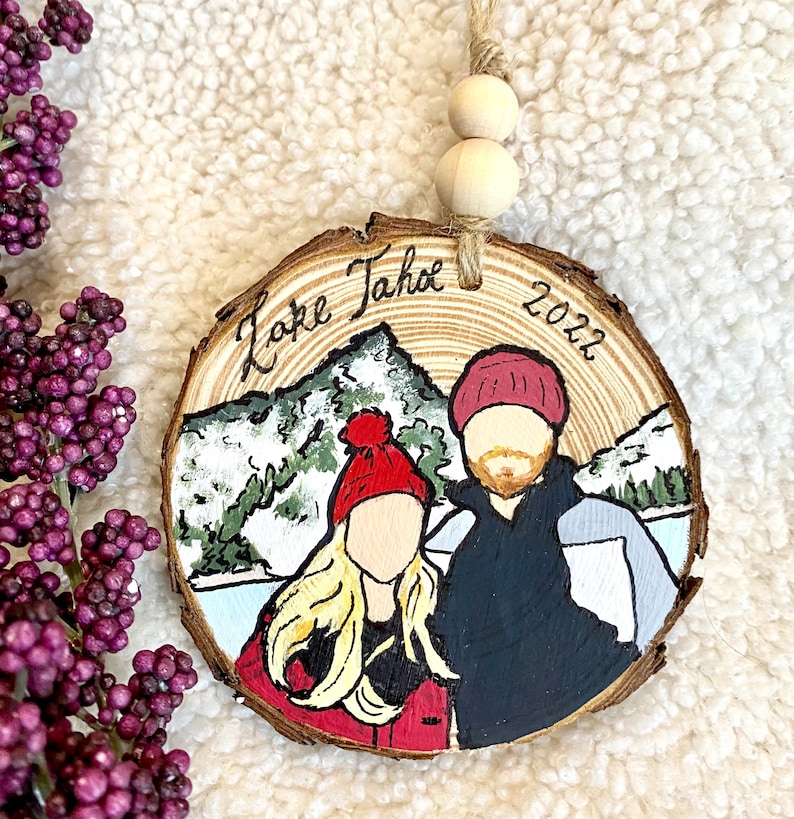 May include: A wooden ornament with a painted scene of a couple in winter clothing standing in front of a mountain with the text "Lake Tahoe 2022" on the wood.