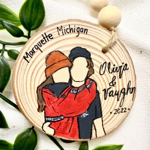 May include: A wooden round ornament with a painted image of a couple hugging. The ornament is decorated with the text "Marquette Michigan" and "Olivia & Vaughn 2022."