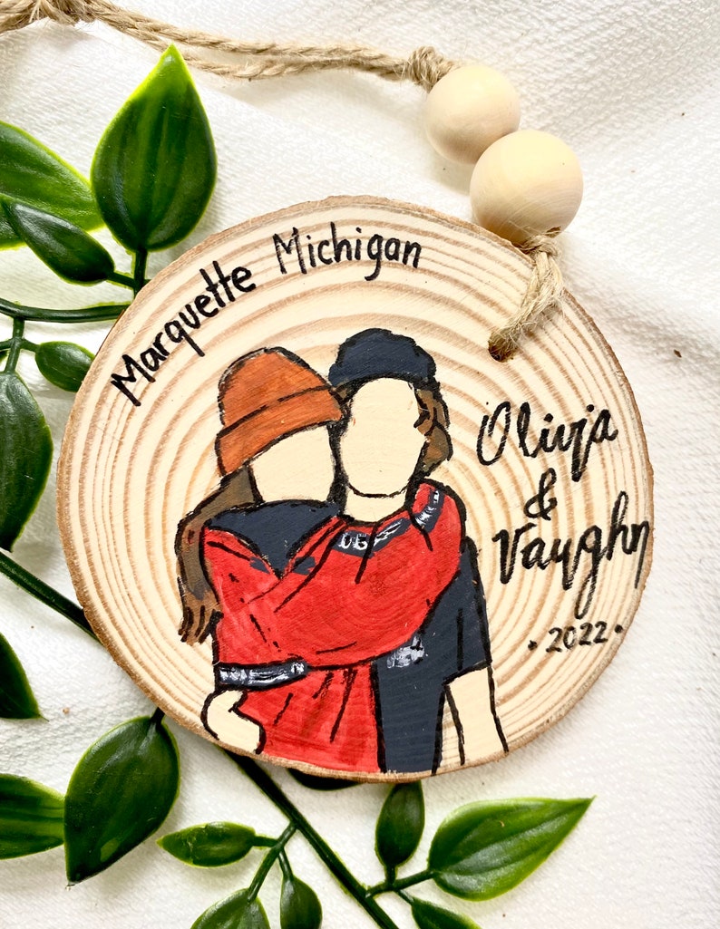 May include: A wooden round ornament with a painted image of a couple hugging. The ornament is decorated with the text "Marquette Michigan" and "Olivia & Vaughn 2022."