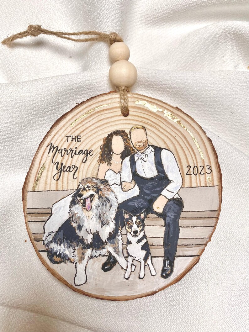May include: A round wooden ornament with a gold accent, featuring a painted portrait of a couple and two dogs. The text "THE marriage Year 2023" is written on the ornament.