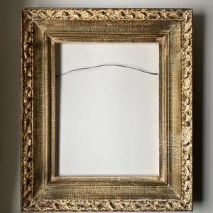 May include: Ornate gold-colored picture frame with a detailed, textured border. The frame has a rectangular shape and a distressed finish, giving it a vintage appearance. The interior is empty, revealing a white backing and a wire for hanging.