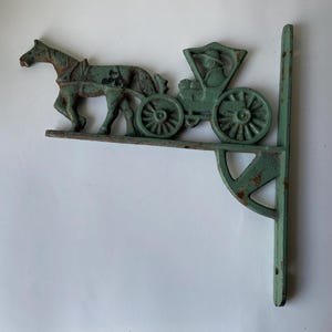 May include: A cast iron decorative bracket in a distressed teal finish. The bracket features a horse-drawn carriage design. The carriage has spoked wheels and a driver. The bracket is designed to be mounted on a wall.