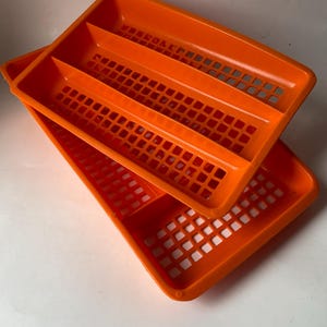 May include: Two bright orange plastic trays. Each tray has a rectangular shape with a grid-like pattern in the center and three divided sections. The trays are stacked on top of each other.