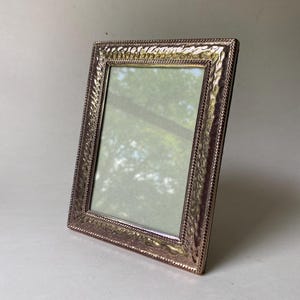May include: A rectangular, rose-gold picture frame with a textured border and a rope-like trim. The frame is angled slightly, revealing a glimpse of a green and white background within.