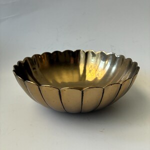 May include: A polished brass bowl with a scalloped edge design. The bowl features a rounded base and vertical indentations along the sides, creating a petal-like appearance with a reflective metallic gold finish throughout the interior and exterior.