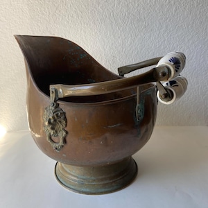 May include: A copper coal scuttle with a lion's head emblem and ceramic handles with blue floral designs. The scuttle has a rounded base and a curved spout. The handles are made of wood and metal.
