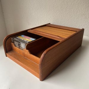 May include: A wooden cassette tape storage box with a sliding top. The box is made of light brown wood and has a curved front. Several cassette tapes are visible inside the box.