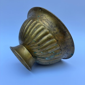 May include: A brass bowl with a fluted, ribbed exterior and a wide, flared rim. The upper portion of the bowl features an engraved floral pattern. The metal surface shows signs of age, including dark patina and small spots of green oxidation.