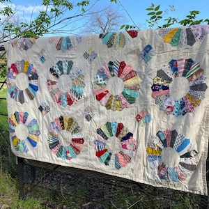 May include: A vintage quilt with a cream-colored background and colorful, circular floral patterns. The quilt is draped over a black metal fence outdoors, with green foliage and a blue sky visible in the background. The floral designs feature various fabric patterns.