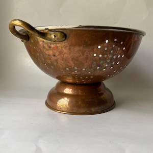 May include: A vintage copper colander with a pedestal base and a single handle. The bowl is perforated with small holes and has a slightly tarnished appearance. The handle is a lighter gold color.