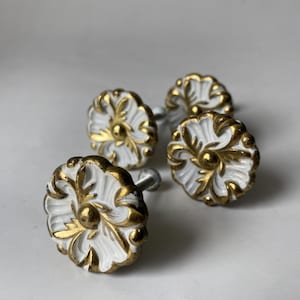 May include: Four white and gold floral cabinet knobs. The knobs have a detailed, ornate design with gold accents and a silver screw. The knobs are arranged on a white surface.