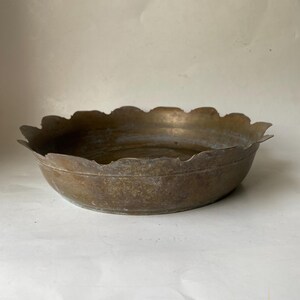 May include: A vintage brass bowl with a scalloped rim. The bowl has a tarnished, aged appearance, with variations in color from light to dark brown. The shallow bowl is likely decorative or for serving.